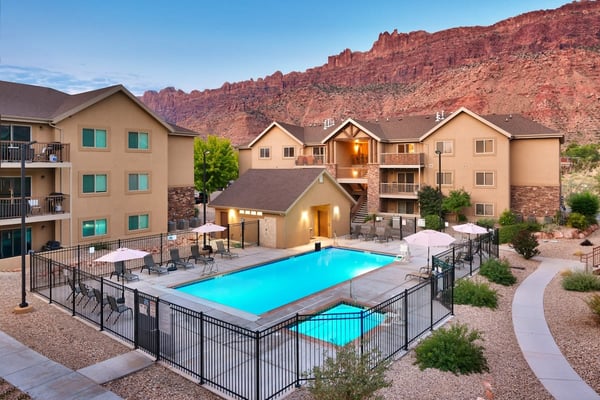 Resort complex with tan multi-story buildings, illuminated pool with black fencing, lounge chairs, and red rock cliffs in background