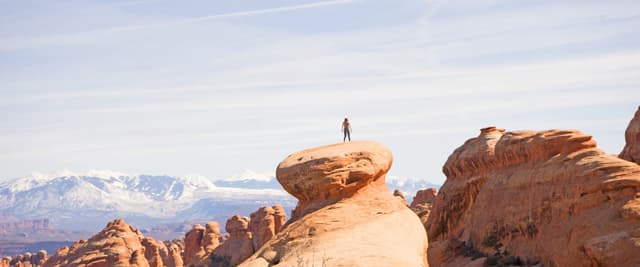 Person standing atop distinctive red rock formation with snow-capped mountains visible in distant background